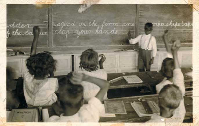 Aboriginal children in a classroom. Some students are raising their hands and one student stands at the front of the classroom in front of a blackboard with cursive chalk writing.