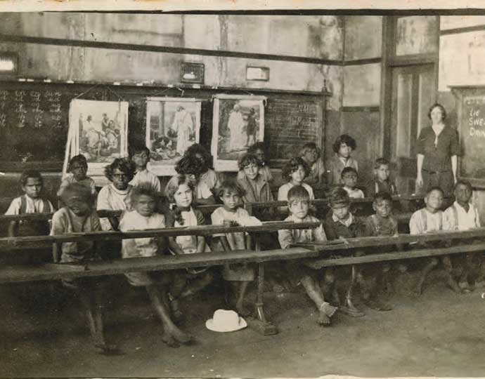 An old black and white photograph of a classroom with the students sitting at desks with the teacher