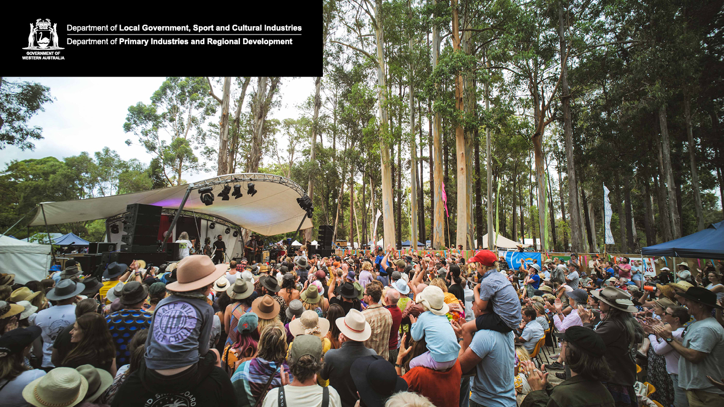 A crowd of people watch and listen to a band on a stage , surrounded by large trees.