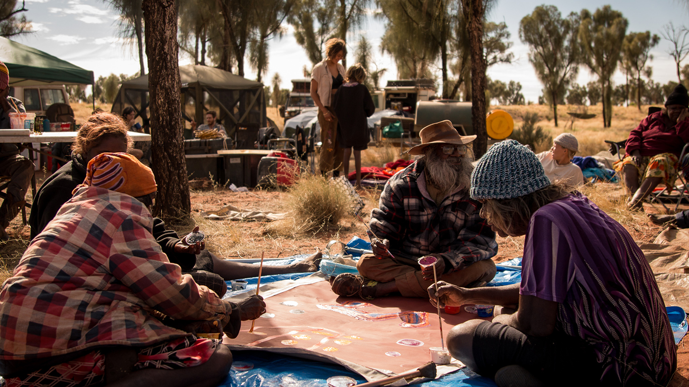 A small group of Aboriginal people sit on the ground painting on an unstretched canvas