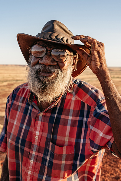 A smiling Helicopter Tjungurrayi wearing a hat standing in the outback. 