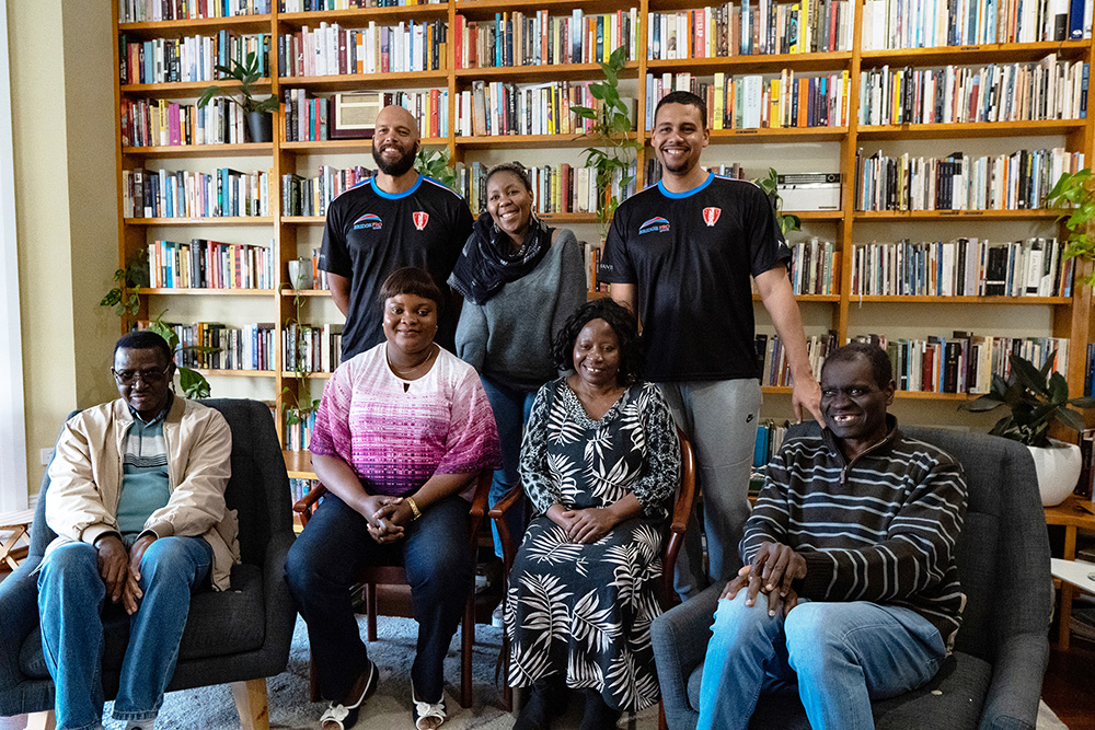 A group of people both sitting on chairs and standing in front of a library of books.