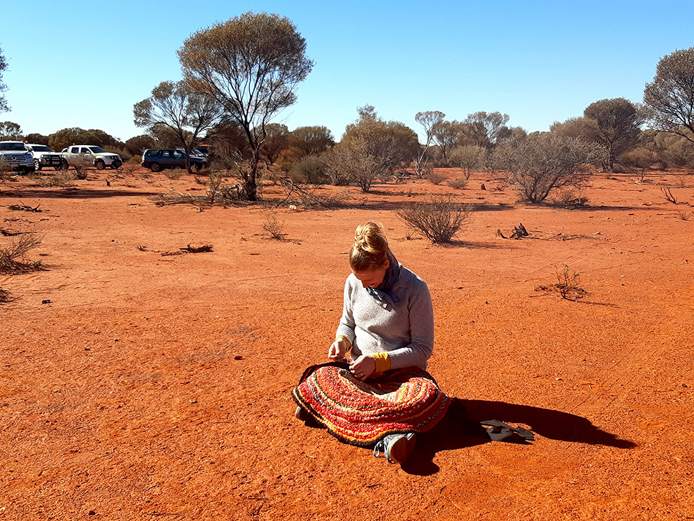 A lady weaving a rug sitting on the desert.