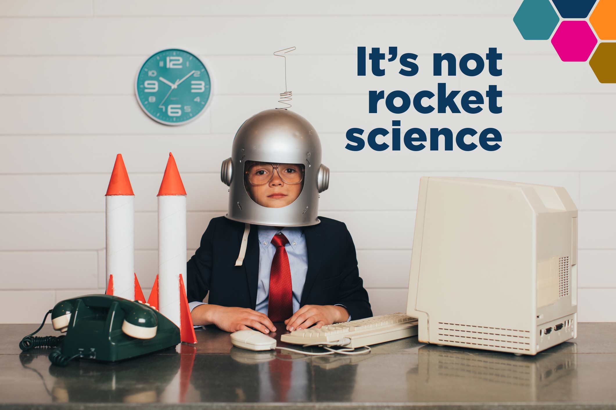 A child dressed in a business suit wearing a pretend space helmet sitting at a desk. The words, ‘It’s not rocket science’ is superimposed.