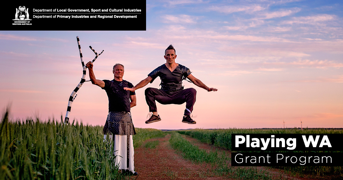 Two dancers in a field, one standing holding a painted branch, the other one jumping in mid-air.