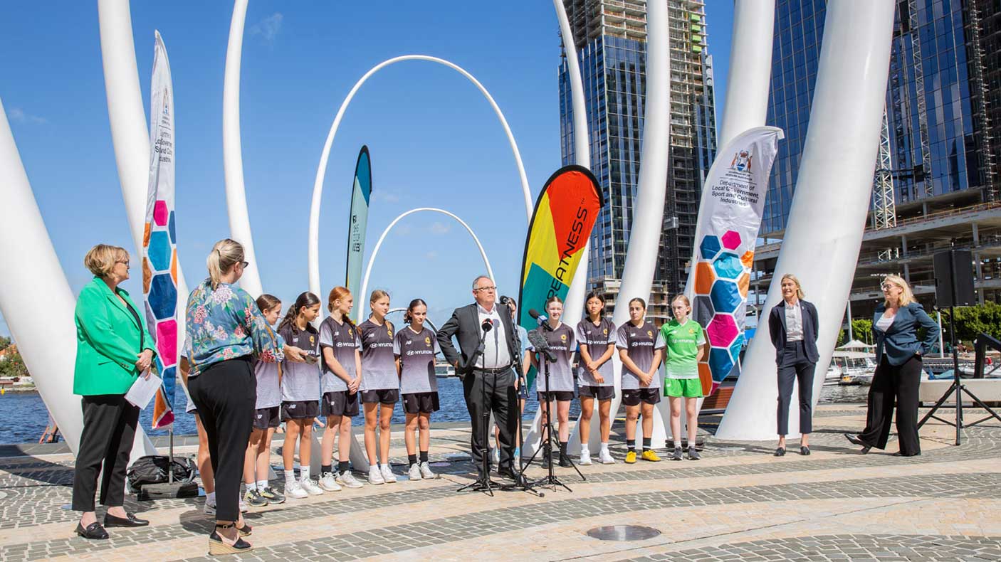 A group of people behind a speaker with banners and a sculpture at Elizabeth Quay, Perth.
