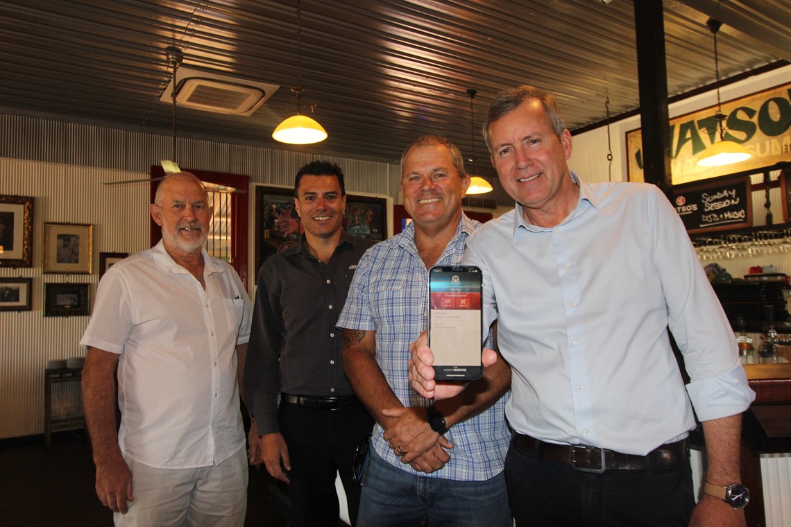 A group of men in a pub, one holding up a smartphone displaying an app