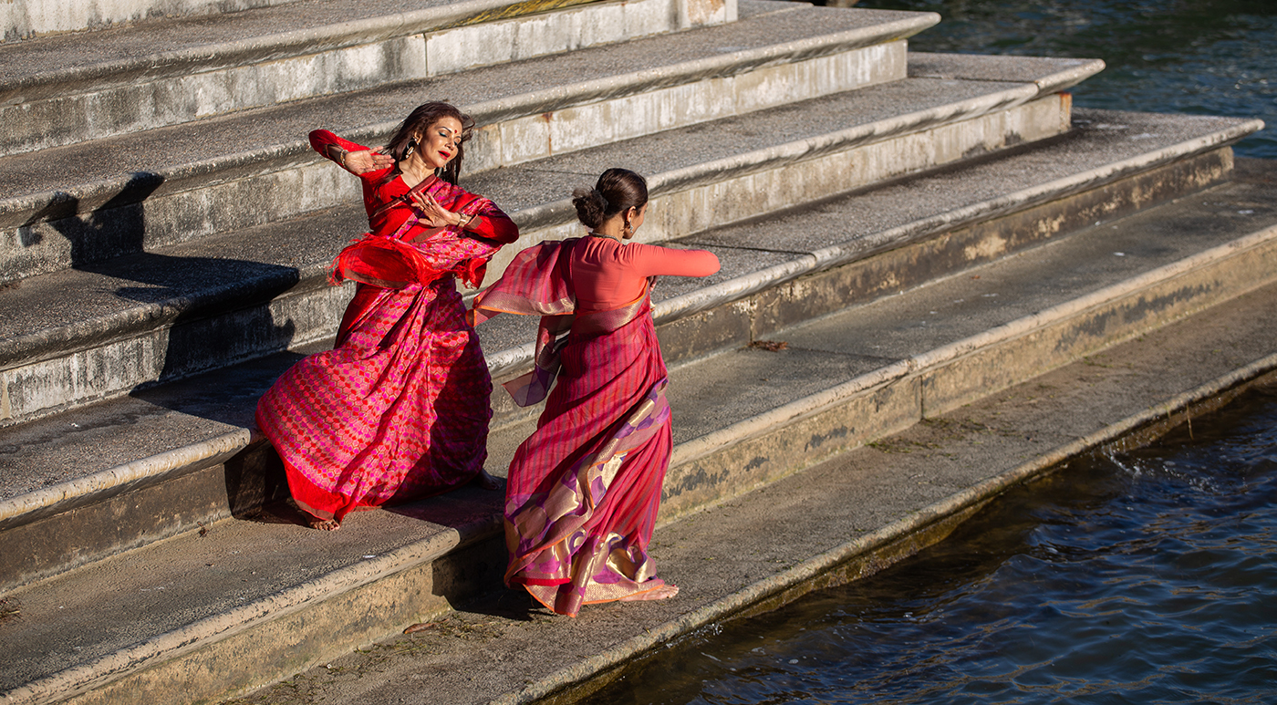 Two Indian female dancers dressed in traditional costumes dancing on the bank of a river.