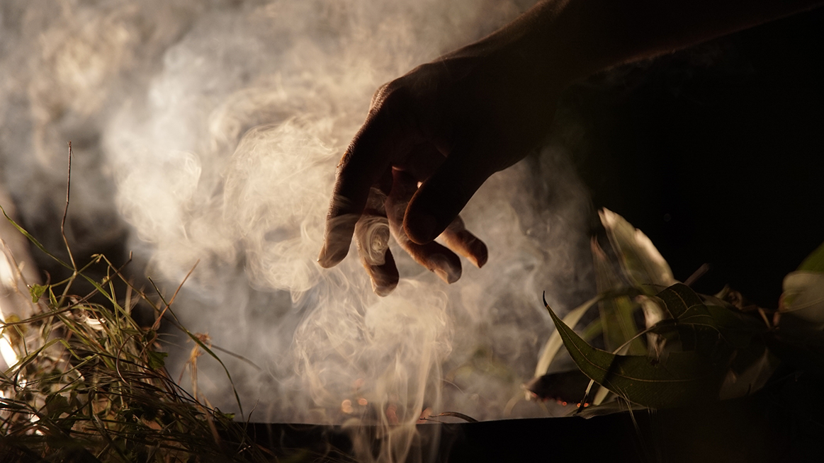 A hand inside smoke emanating from a smoking ceremony container burning bush plants.