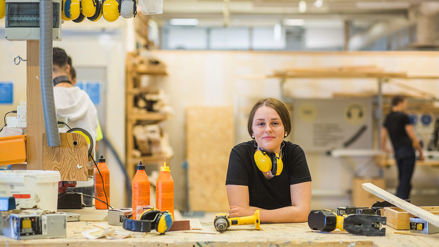 Portrait smiling young female carpentry trainee leaning on workbench with power tools at illuminated workshop - stock photo