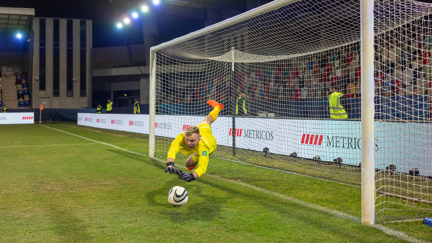 In a stadium, a soccer goalie leaps out of the goals to deflect a incoming soccer ball.