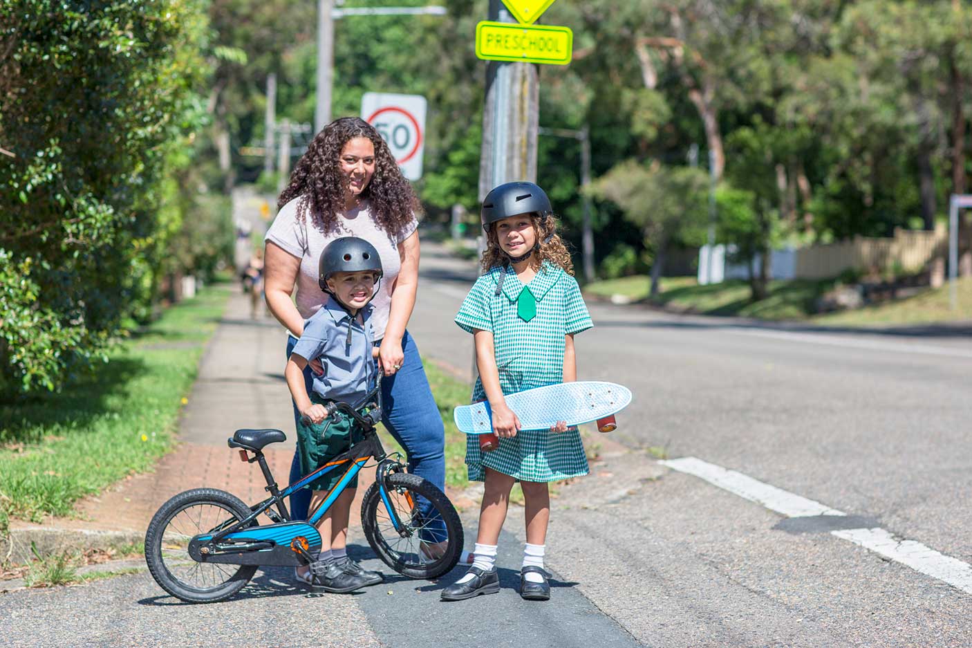 A mother with her school-aged children going to school. ONe child has a bike and the other a skateboard.