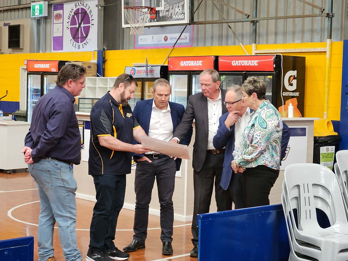 A group of 6 people pointing at a piece of paper. They are inside a basketball stadium.