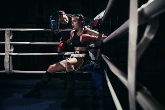 Photo of a female boxer sitting in a boxing ring corner. She is listening to advice from her trainer.