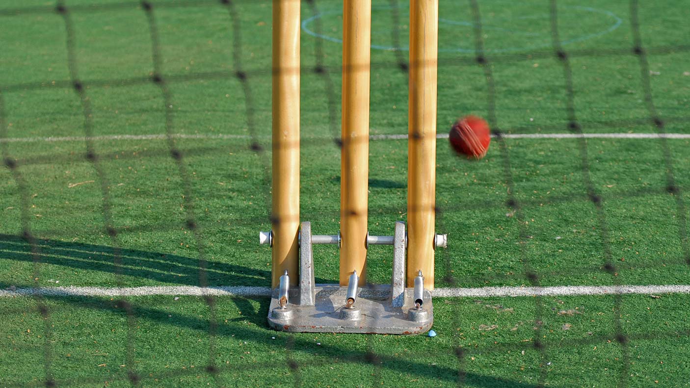 Close-up of the wickets and red cricket ball on green pitch.
