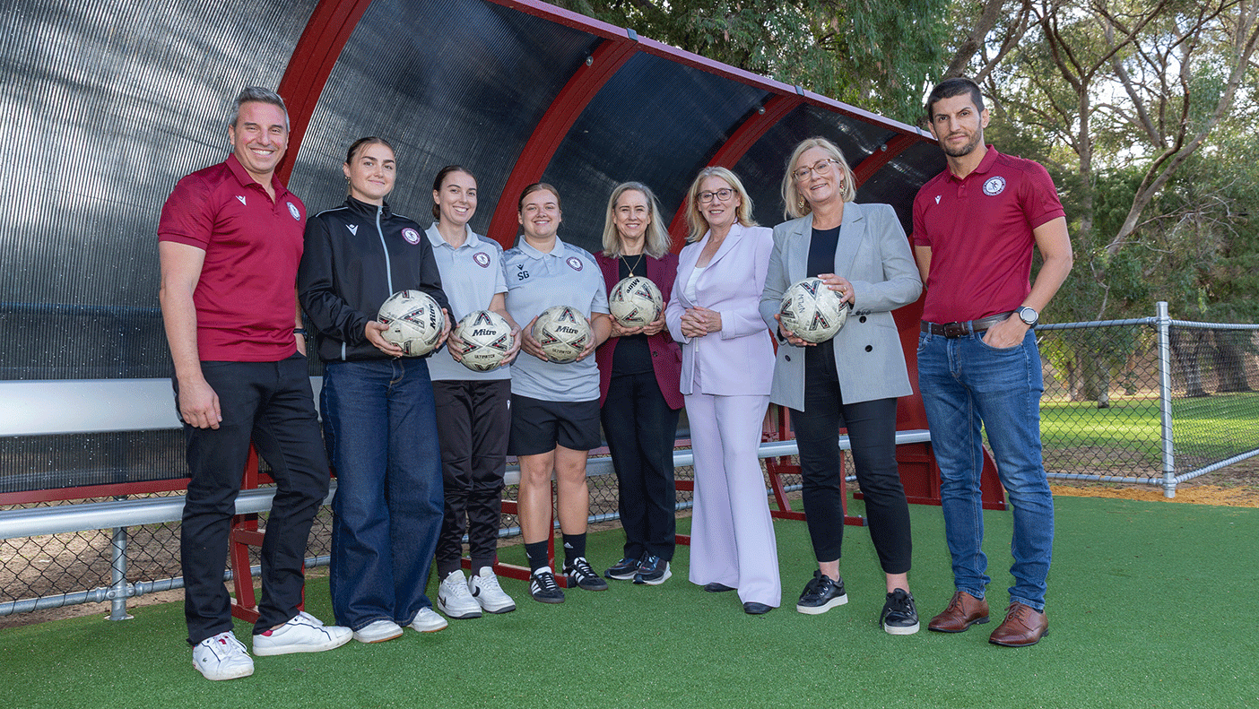 A group of people holding soccer balls stand under a shelter on a football pitch